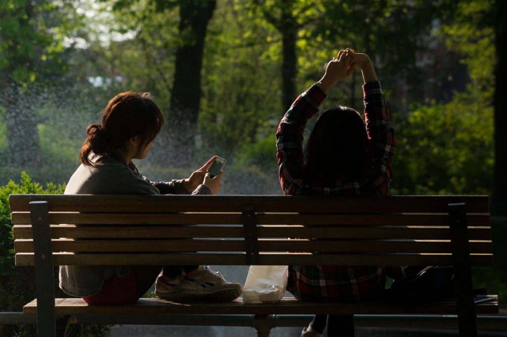 Rest for Your Soul_two women on park bench, one looking at phone, one stretching arms overhead