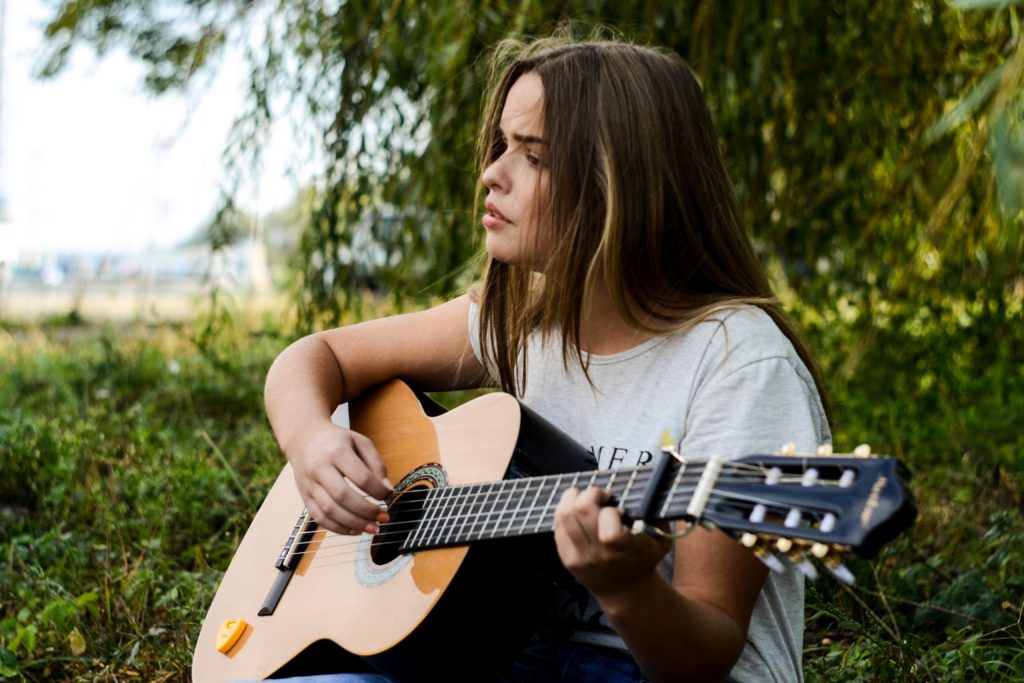 An Unparalleled Way to Clear Your Mind to Hear from God_young woman sitting outdoors, strumming guitar, green of tree behind her