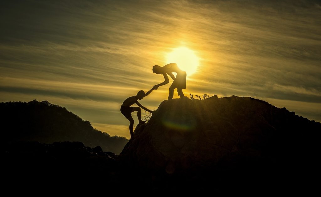 Silhouette of teen boy on top of a mountain or plateau reaching with one hand to pull another teen boy up the mountain