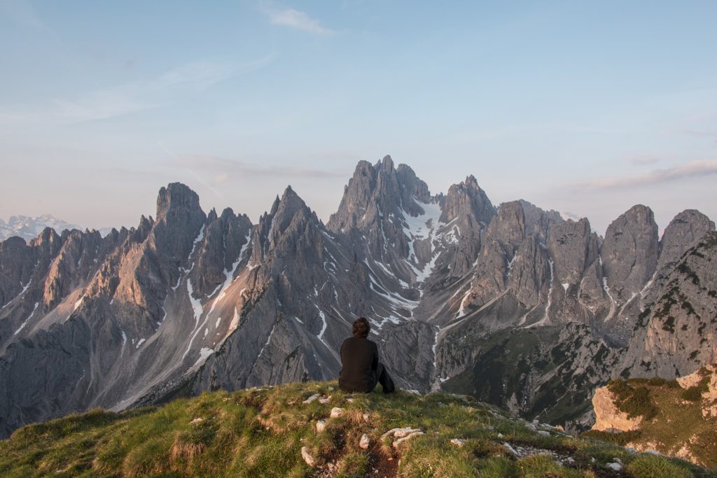 View from back of man in black jacket, sitting on green ridge, looking at rocky, craggy mountains