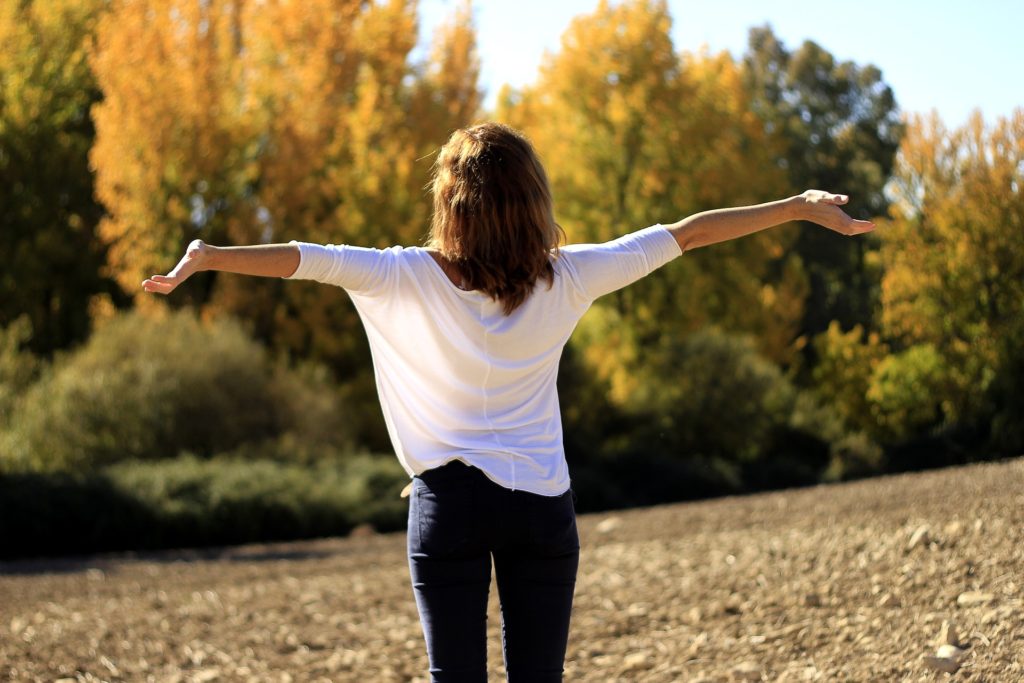 Woman in white blouse, blue jeans, facing away from camera, arms outstretched, looking at trees, sun shining