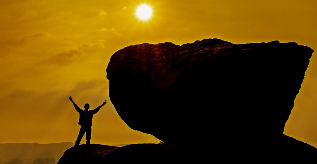 Silhouette of man with arms raised, looking at sun, standing next to enormous rock, double his height