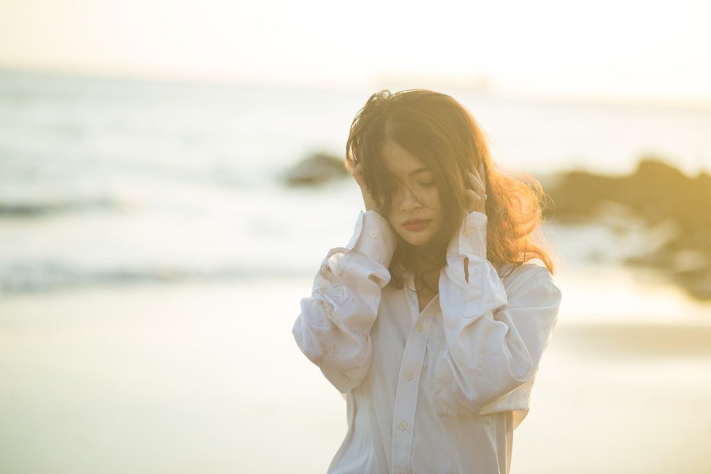 Woman in long-sleeved blouse walking by ocean, hands to head, eyes downcast, sad