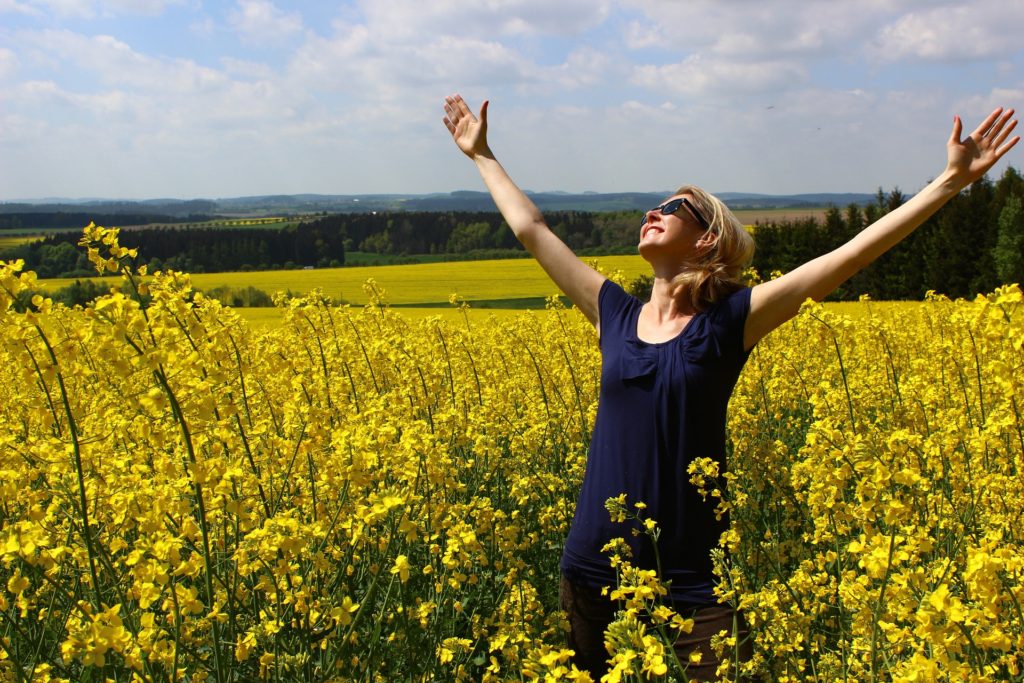 Woman in navy blue blouse, standing in field of yellow flowers, arms raised, grateful smile on her face